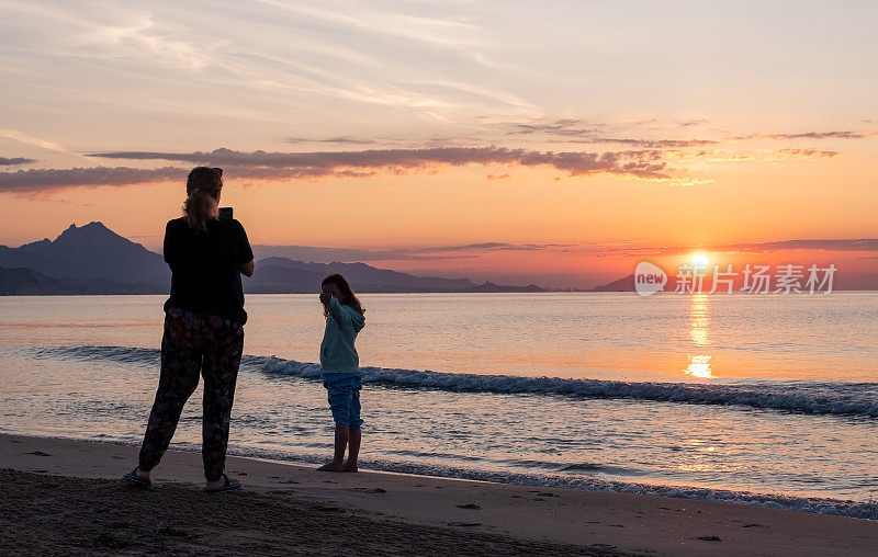 海面上的日出。在海滩上，一位年轻的妇女和七岁的女孩正在看日出。一位女士用智能手机给孩子拍照。