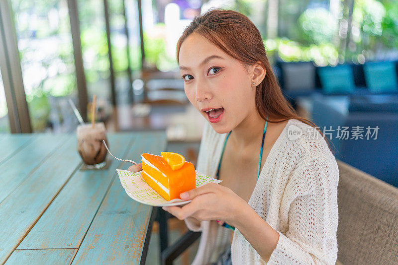 年轻的亚洲女子在海滩咖啡馆喝着冰拿铁，一边享用着一片橘子蛋糕。穿着沙滩装的女性数字游牧民在度假氛围中休息。穿着沙滩装的亚洲女人在咖啡馆里放松。