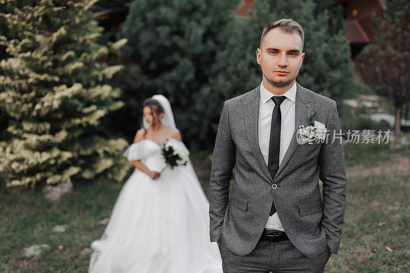 新娘和新郎在大自然中的肖像。新郎在前景，新娘站在他身后，穿着白色宽松的礼服，拿着一束花，低头看着。时尚的新郎
