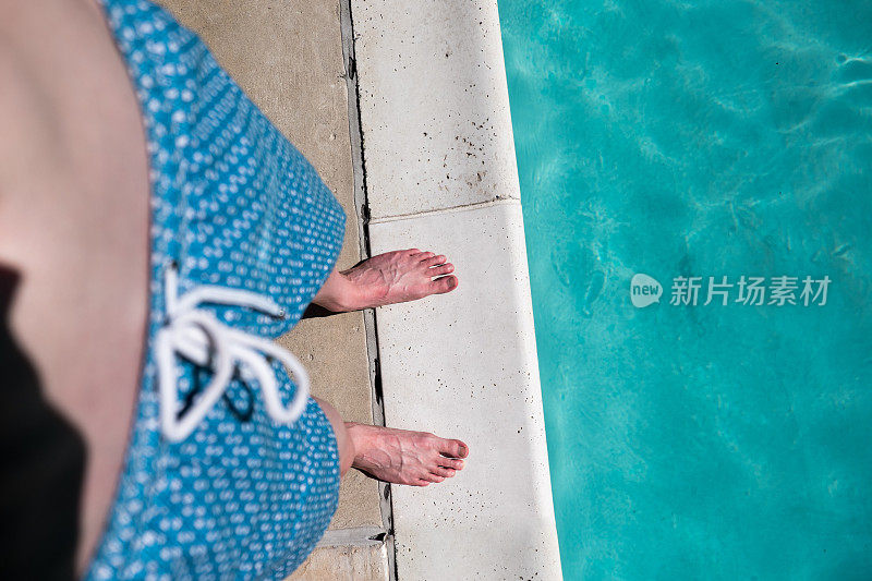 镜头下，一个男人赤裸着脚，穿着蓝色泳裤站在一个室外游泳池的边上。一个穿着泳衣的白人男子站在泳池边。