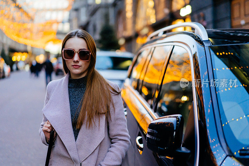 美丽年轻的白人女人站在市中心的豪华越野车旁。时尚的女模特戴着太阳镜拍照附近昂贵的汽车。