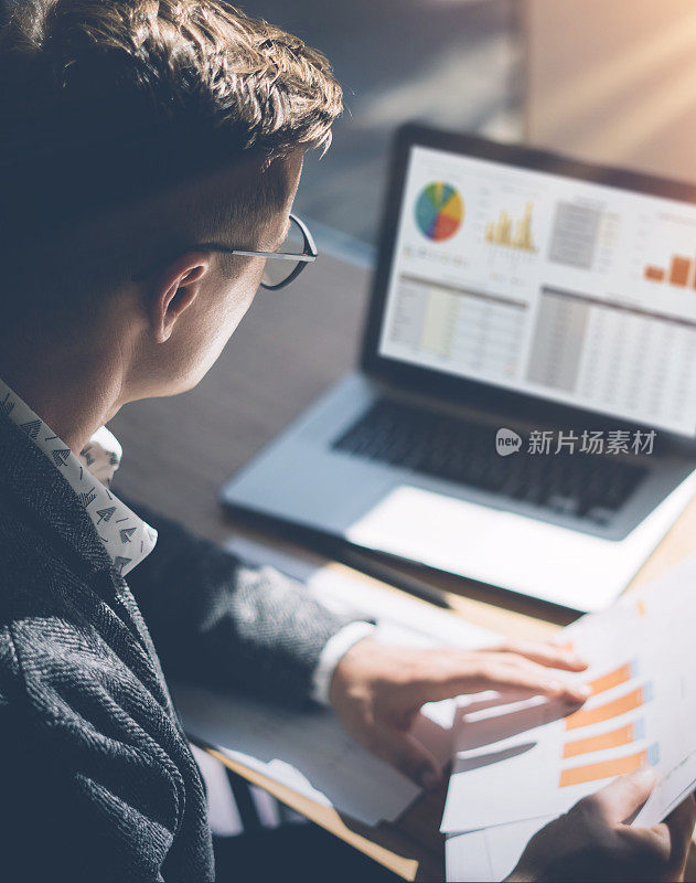 年轻的金融市场分析师戴着眼镜，坐在阳光明媚的办公室里，用笔记本电脑工作。商人分析手中的文件。图形和图表在笔记本屏幕上。垂直。