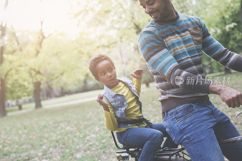 微笑的非裔美国父亲驾驶他的小女孩在自行车槽公园。