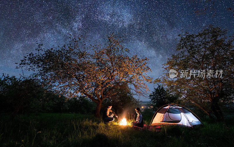 浪漫的情侣游客坐在篝火附近的帐篷下的树和美丽的夜空满是星星和银河。晚上露营