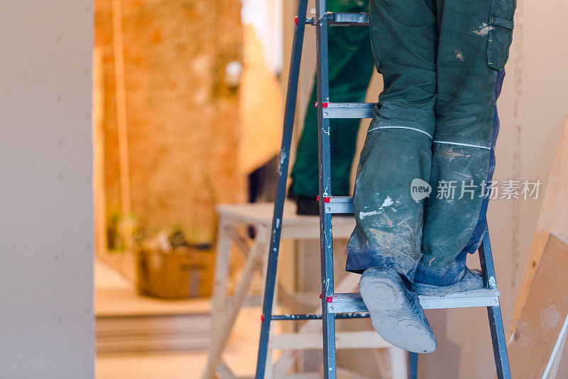 梯子上穿着绿色工作服的工人的腿。石膏板干墙工人为公寓内的石膏板墙正在施工、改造、翻新、修复和重建中。