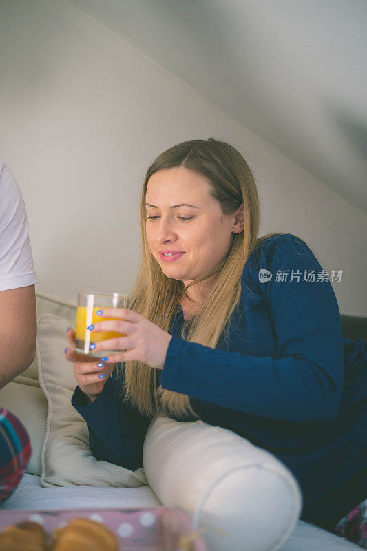 一个快乐的年轻女人正躺在床上准备吃早餐。拿着一杯橙汁