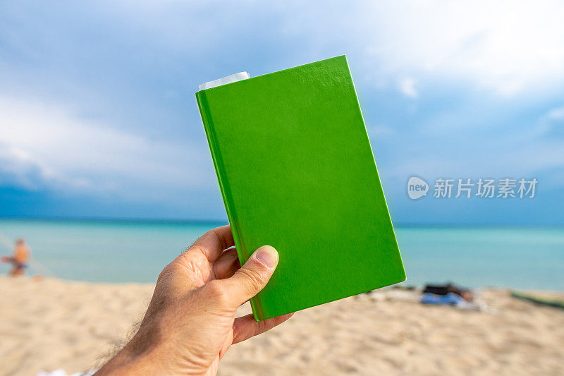 手拿一本绿色的书在一个平静和阳光的海滩前