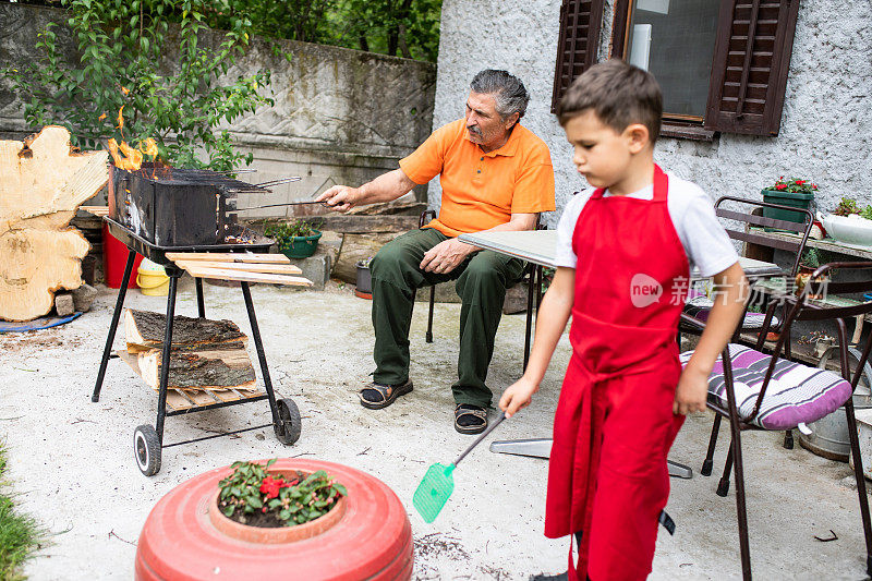 一个可爱的小男孩在玩苍蝇拍，而他的爷爷正在点烧烤