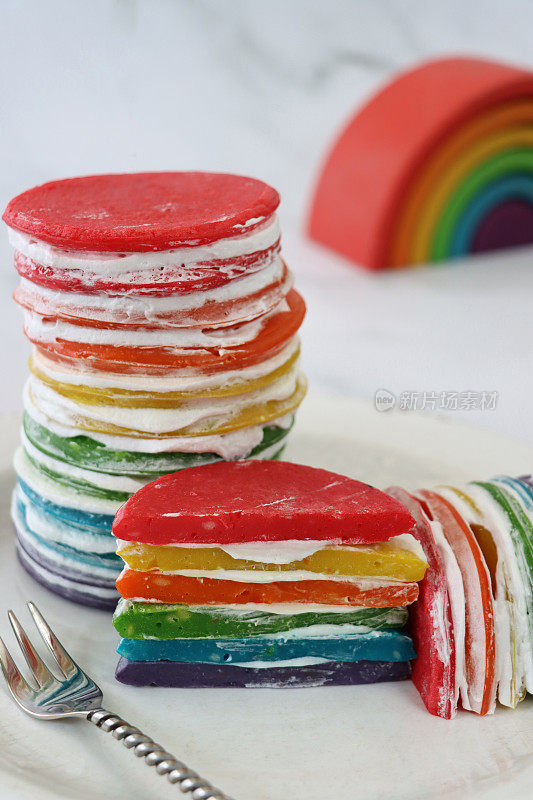 白色的盘子上有一堆彩虹色的煎饼和半切的薄饼，一层层的奶油煎饼，旁边是彩虹装饰的大理石效果的背景，聚焦前景