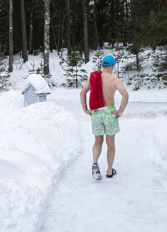 一名身穿泳裤和沙滩浴巾的白人男子沿着积雪覆盖的乡村道路去蒸桑拿。桑拿和芬兰传统。