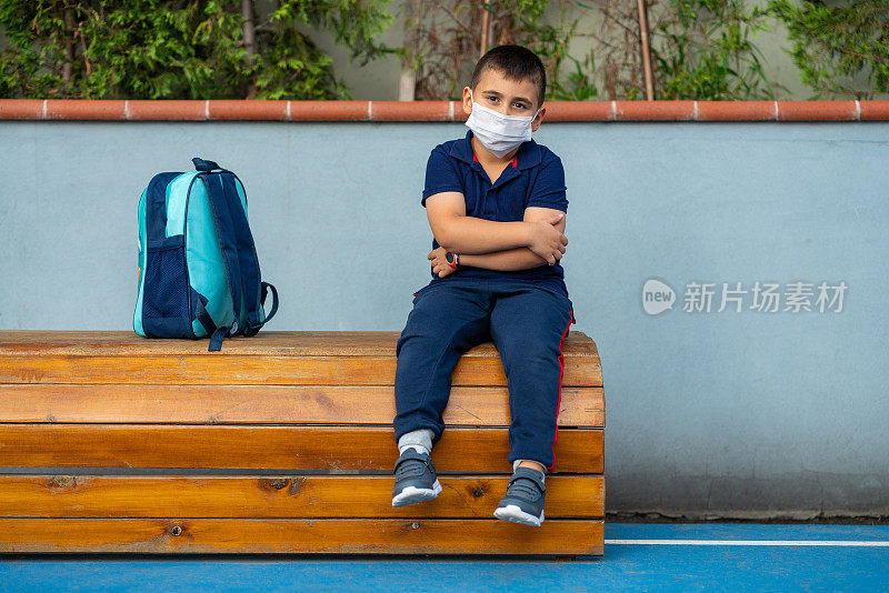 小男生戴着防护口罩，坐在学校前面的长凳上沉思