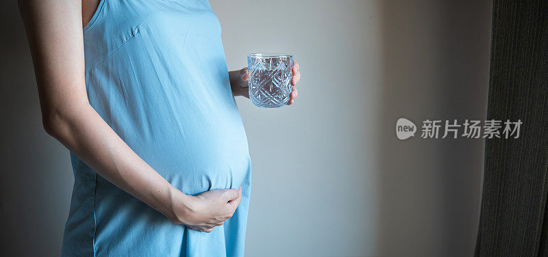 一个孕妇抱着她的肚子和一杯水在卧室靠墙