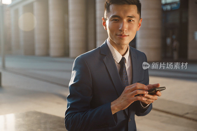 一位身穿蓝色西装的亚洲商人在美丽的晚霞中手持智能手机，看着镜头