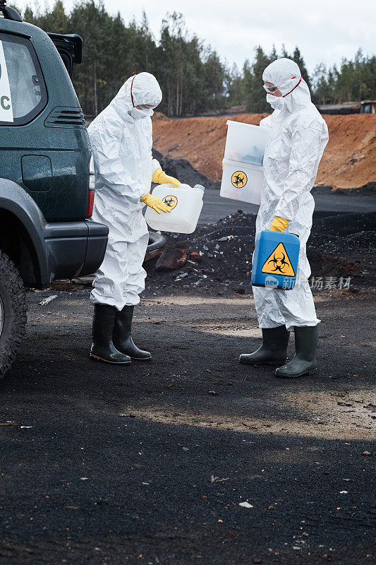 两名穿着防护工作服和胶靴的生态学家站在被污染的土地上，拿着装有有毒物质的容器