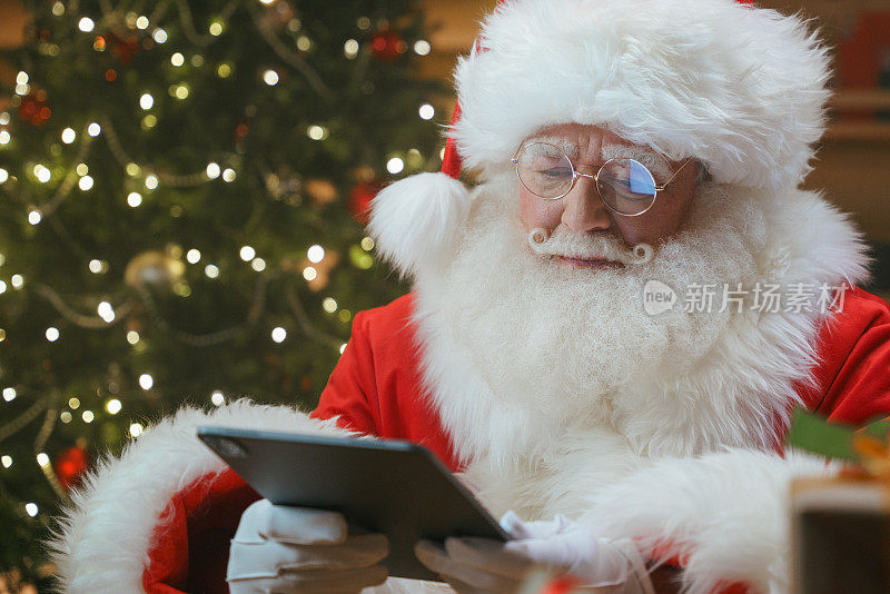 这是一个圣诞老人聚精会神地看着数码平板电脑的特写镜头，他坐在装饰有圣诞树的小木屋里的一张桌子前