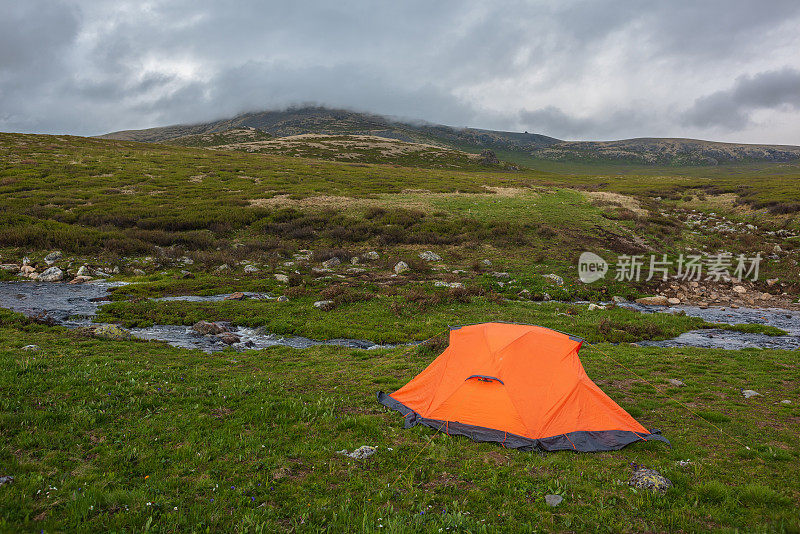 橙色帐篷靠近清澈的小溪，在低垂的雨云中可以看到山脉。引人注目的高山景观，山溪和绿色的山丘在铅灰色的低云。冻土带阴森森的景象