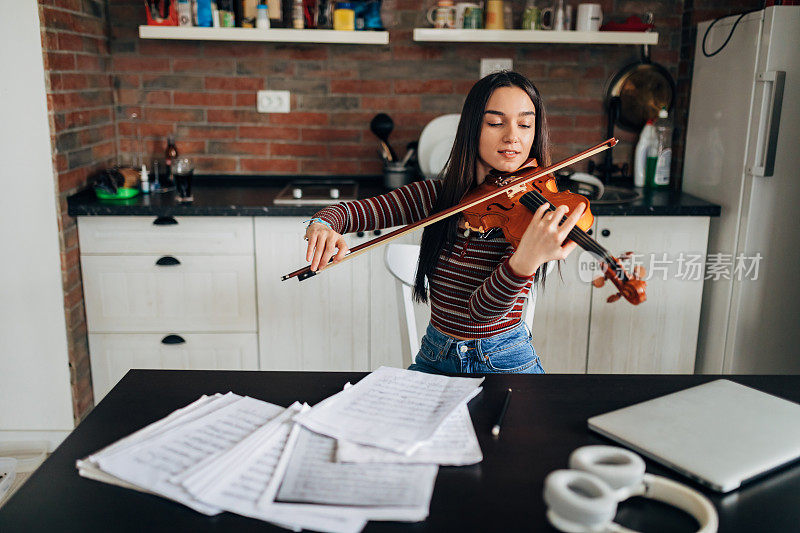 年轻的女小提琴手坐在餐厅的桌子旁，边看乐谱边拉小提琴。