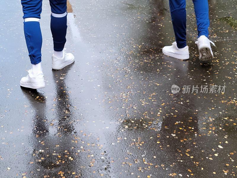两个穿着白色运动鞋和蓝色运动裤的男人走在潮湿的人行道上。