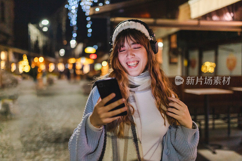 在城市的夜晚，微笑的年轻女子用她的智能手机听音乐