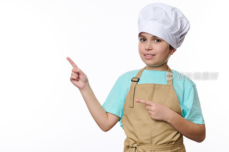 戴着厨师帽和围裙的可爱少年指着白色背景上的宣传文字的空余空间，看着镜头