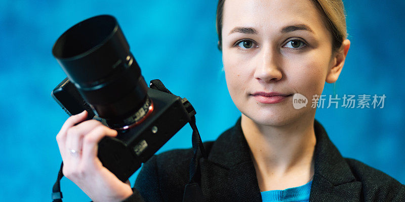 女性肖像的专业摄影师与无反光镜数码相机在工作室与蓝色背景。女士穿着商务黑色夹克和蓝色t恤，室内生活方式概念摄影。