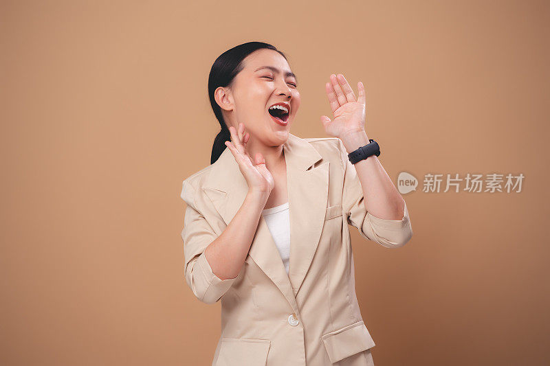 一个亚洲女人开心地微笑着，大声地说着，分享着新闻，独自站在米色的背景上。