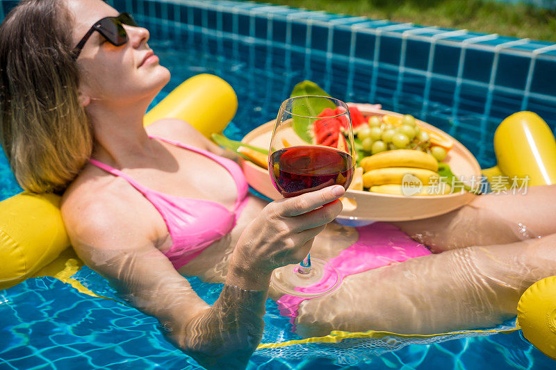 美丽的女人躺在漂浮的吊床上，在游泳池和酒和水果托盘