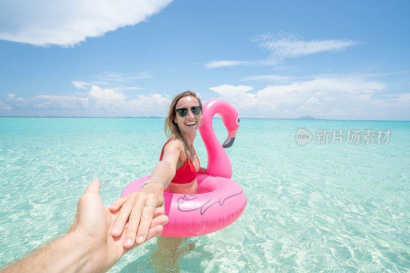 跟随我的概念女人领先的男朋友在田园诗般的海滩充气火烈鸟玩在纯净的水在泰国的岛屿。人们旅行奢侈，乐趣和酷的态度概念