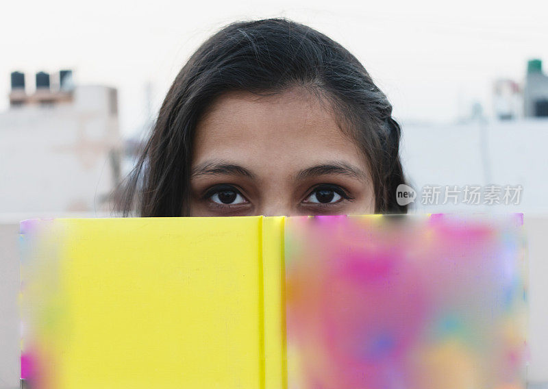 美丽的亚洲女孩用书遮住她的脸，看着相机