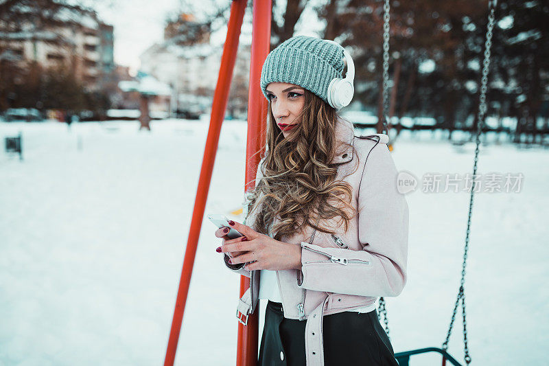 在大雪纷飞的冬日里，用耳机和智能手机的街头时尚女子