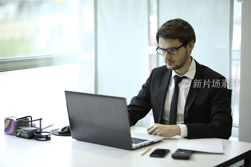 严肃的商人在笔记本电脑上参考文件输入信息。商人在办公室用笔记本电脑计算财务数据。在办公室工作的男人。