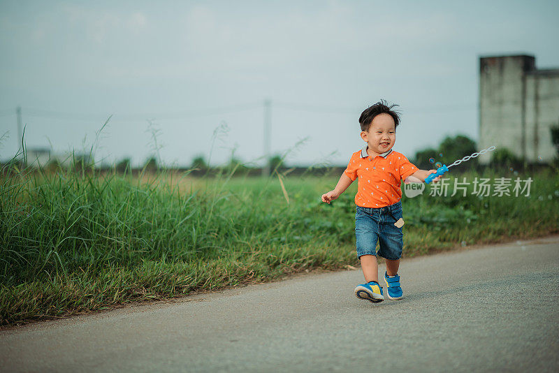 亚洲华人微笑男孩花闲暇时间，手拿泡泡棒，在村里的街道上跑步