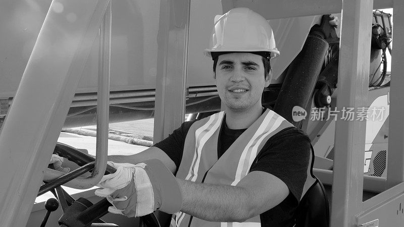 一名年轻男性建筑工人微笑着看着镜头，手里拿着一辆大型叉车的方向盘