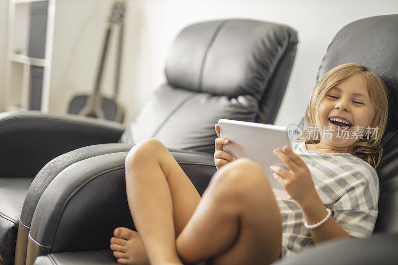 微笑的小女孩穿着睡衣，坐在扶手椅上，在家里使用数码平板电脑