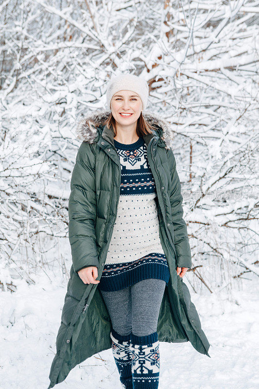 在冬天下雪的日子里，妇女穿着绿色的蓝色皮夹克，民族图案的羊毛连衣裙和帽子走在户外的公园森林里。快乐的独自旅行者探索当地的远足路线。