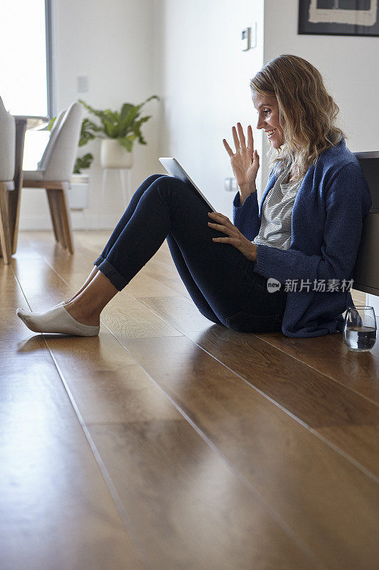 一个愉快的女人一边挥手一边打视频电话坐在家里的地板上