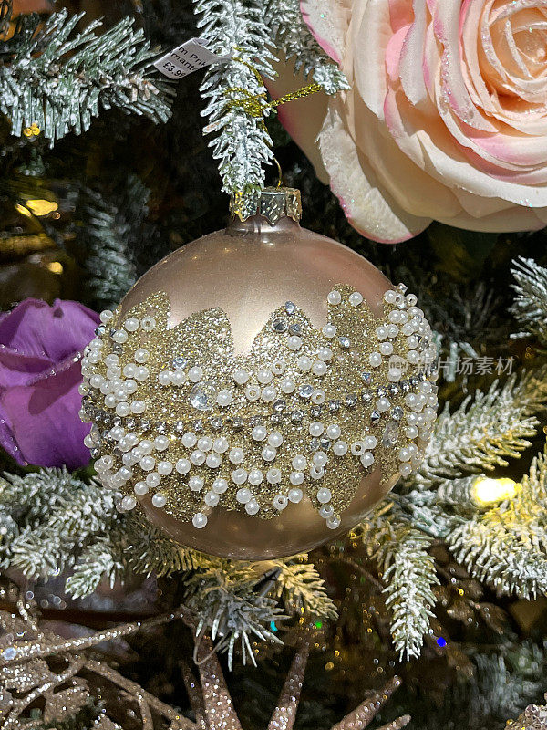 图像单个桃色的小饰物装饰着白色的珠子和金色的闪光，人造的，玫瑰花头饰品挂在人造圣诞树的树枝上，云杉针覆盖着人造的喷雪，模糊的白色精灵灯