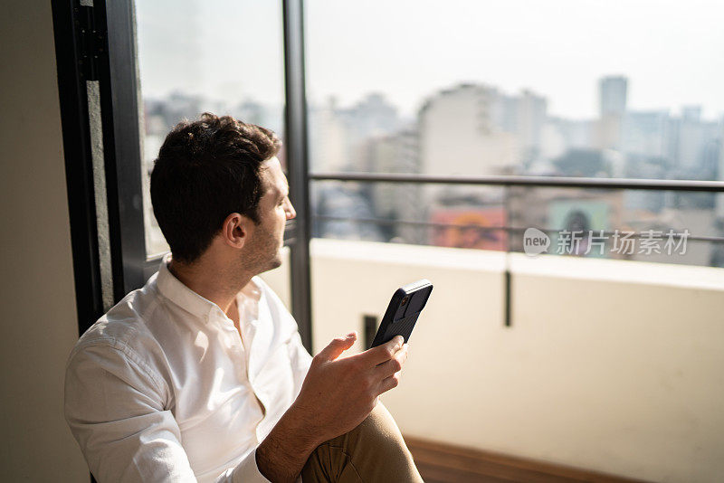 年轻男子坐在窗台上沉思，在家使用智能手机