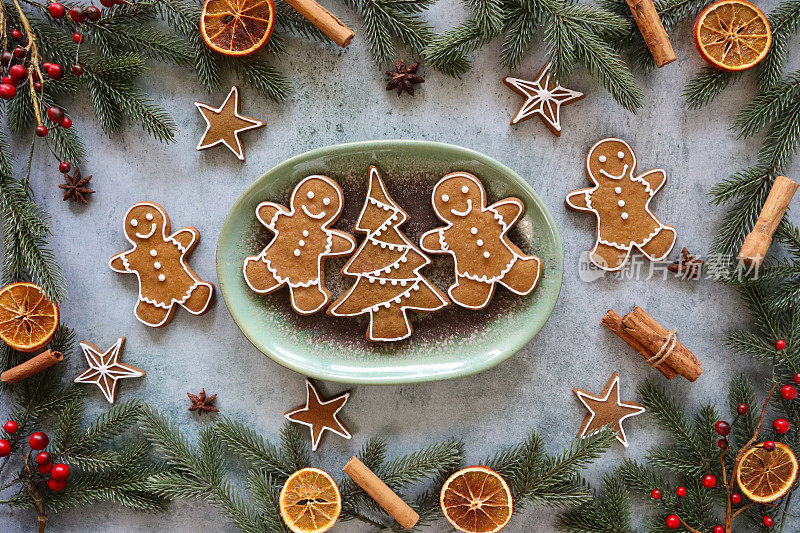 自制的姜饼人和圣诞树形状的饼干的特写图像，这些饼干在椭圆形的盘子里用白色的皇家糖霜覆盖，周围是云杉针、红莓、肉桂棒、八角和柑橘干水果片