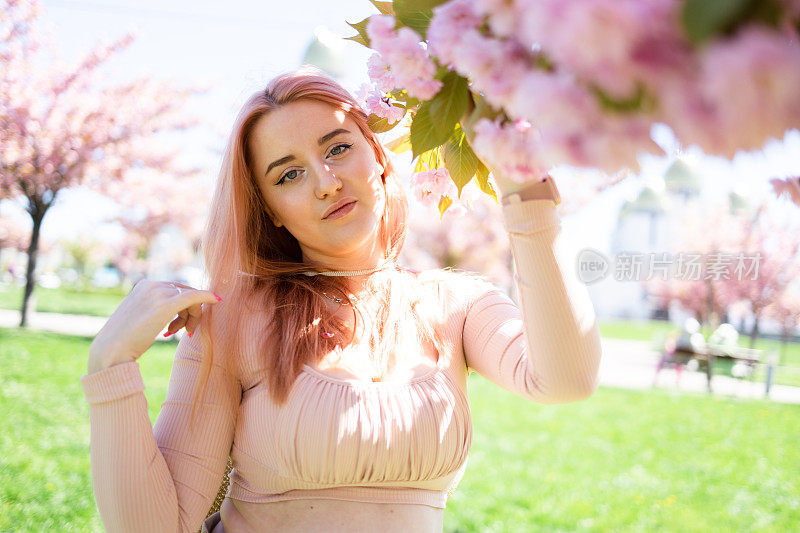 美丽的女孩在樱花树旁。女人穿着裙子和时髦的外套粉红色的花朵盛开。在开花。春天的时间。放松和幸福的概念。