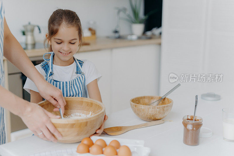 微笑的小女孩助手端着大碗，看着妈妈如何用面粉拌鸡蛋，用巧克力准备美味的蛋糕，在温馨的家庭厨房内部摆姿势，为所有的家庭准备食物