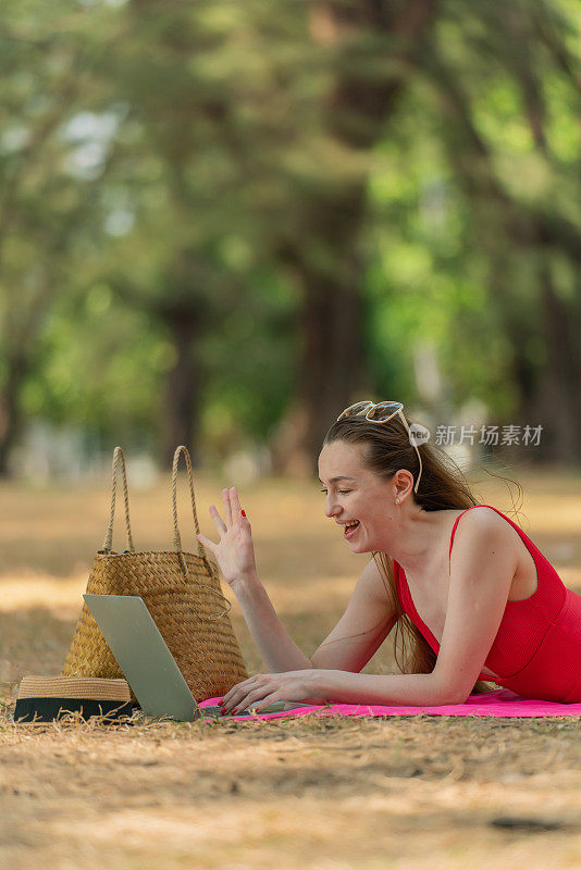 美丽的白人女性性感的女人在红色泳装比基尼戴着太阳镜躺在地上工作在她的电脑笔记本电脑放松在树林里的海滩