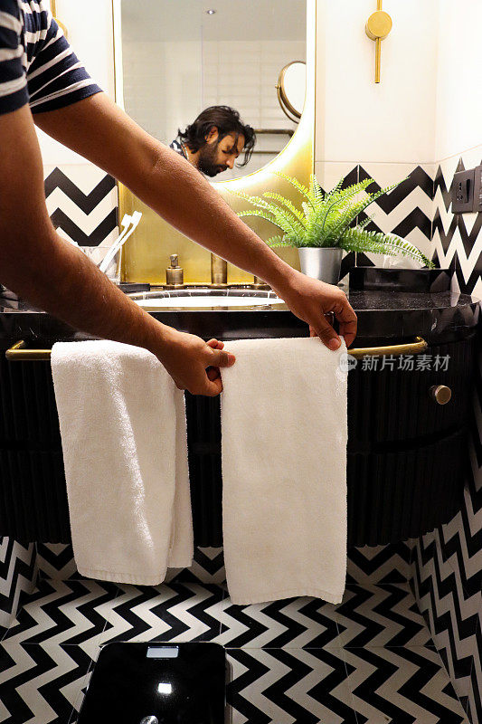 印度男子站在浴室的水槽边，把折叠的白色毛巾挂在毛巾架上，挂在水槽梳妆台上，镜子里的倒影，大理石浴室柜台上金属锅里的人造蕨类植物，称重秤，专注于前景