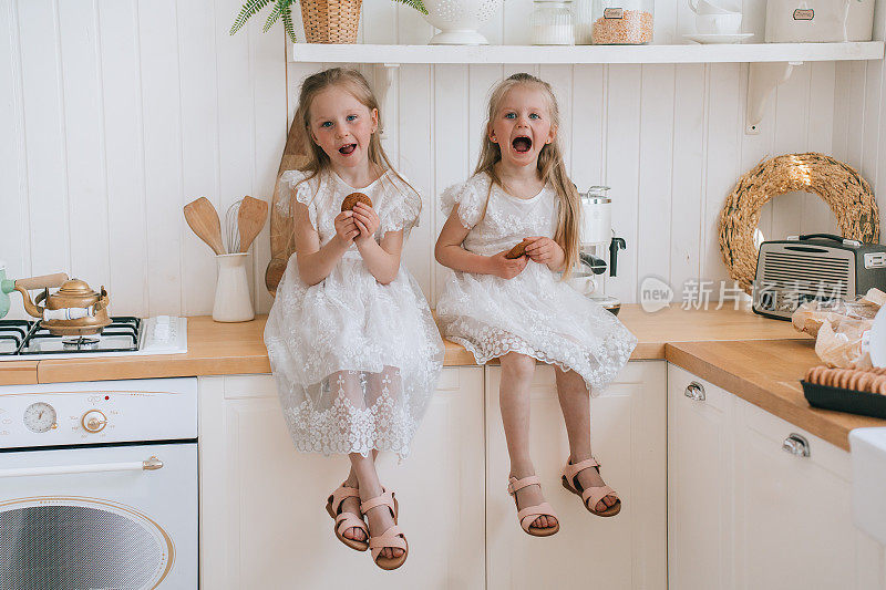 两个小姐妹坐在厨房的桌子上，唱歌，玩，吃饼干。白人小女孩早上在家里吃早餐。姐妹情谊，家庭生活。