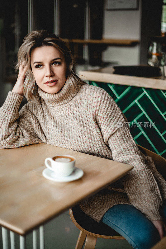 贴心的女人在舒适的咖啡馆喝咖啡。一个穿毛衣的女人坐在咖啡桌旁，手放在头上，沉思着，面前放着一杯咖啡。