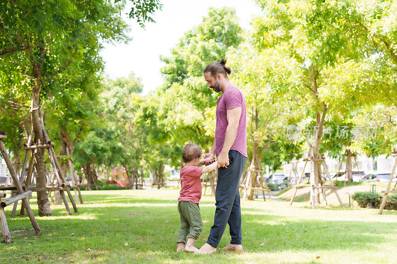 白种人爸爸妈妈牵着他们的小儿子的手，牵着每个孩子的手举起来，爸爸妈妈带着他们可爱的儿子在野餐公园散步，阳光明媚的日子里，三脚碰着草地。