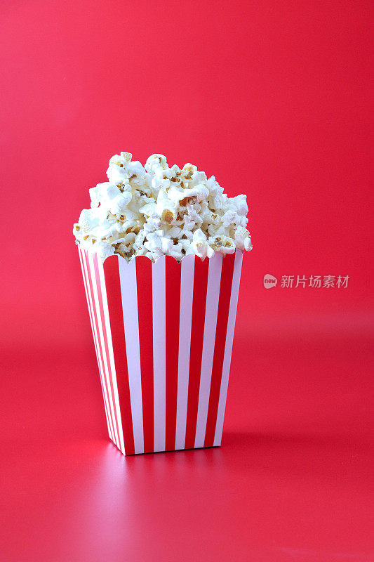 图像的红白条纹纸箱装满爆米花，爆玉米粒，电影休闲食品桶，红色的背景，重点在前景