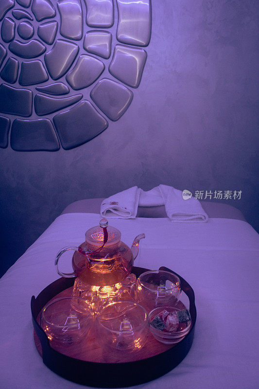 按摩椅上的玻璃茶壶里放着花草茶和蜡烛。水疗沙龙内部。奢侈的生活方式。热中心。茶壶与杯托盘在紫色的光。