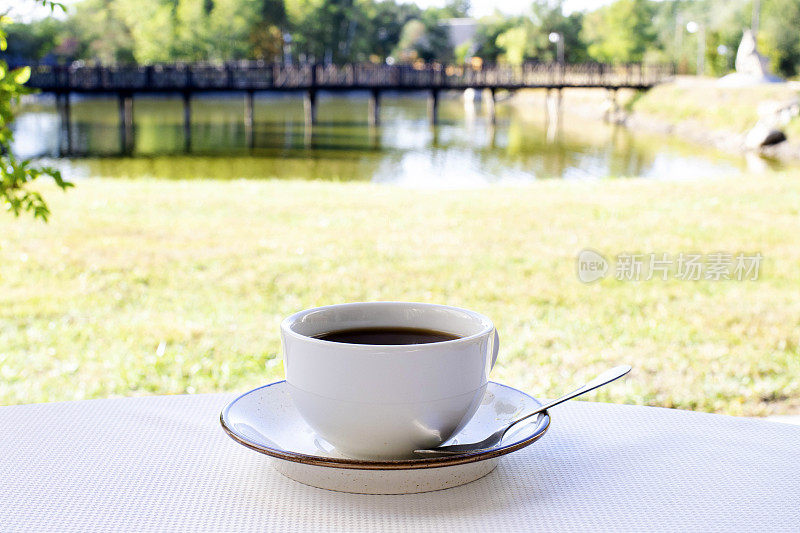 桌子上有一杯白色的咖啡，以河为背景。本质上的茶党。早上喝咖啡