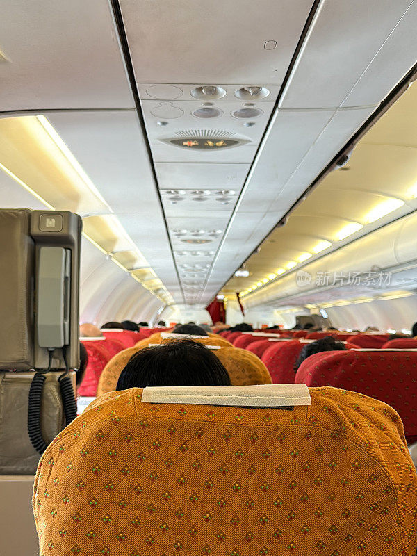 客机经济舱顶置通风控制台特写图像，照明的禁止吸烟和系好安全带标志，乘务员呼叫按钮，后排乘客座椅，缩小视角，聚焦于前景